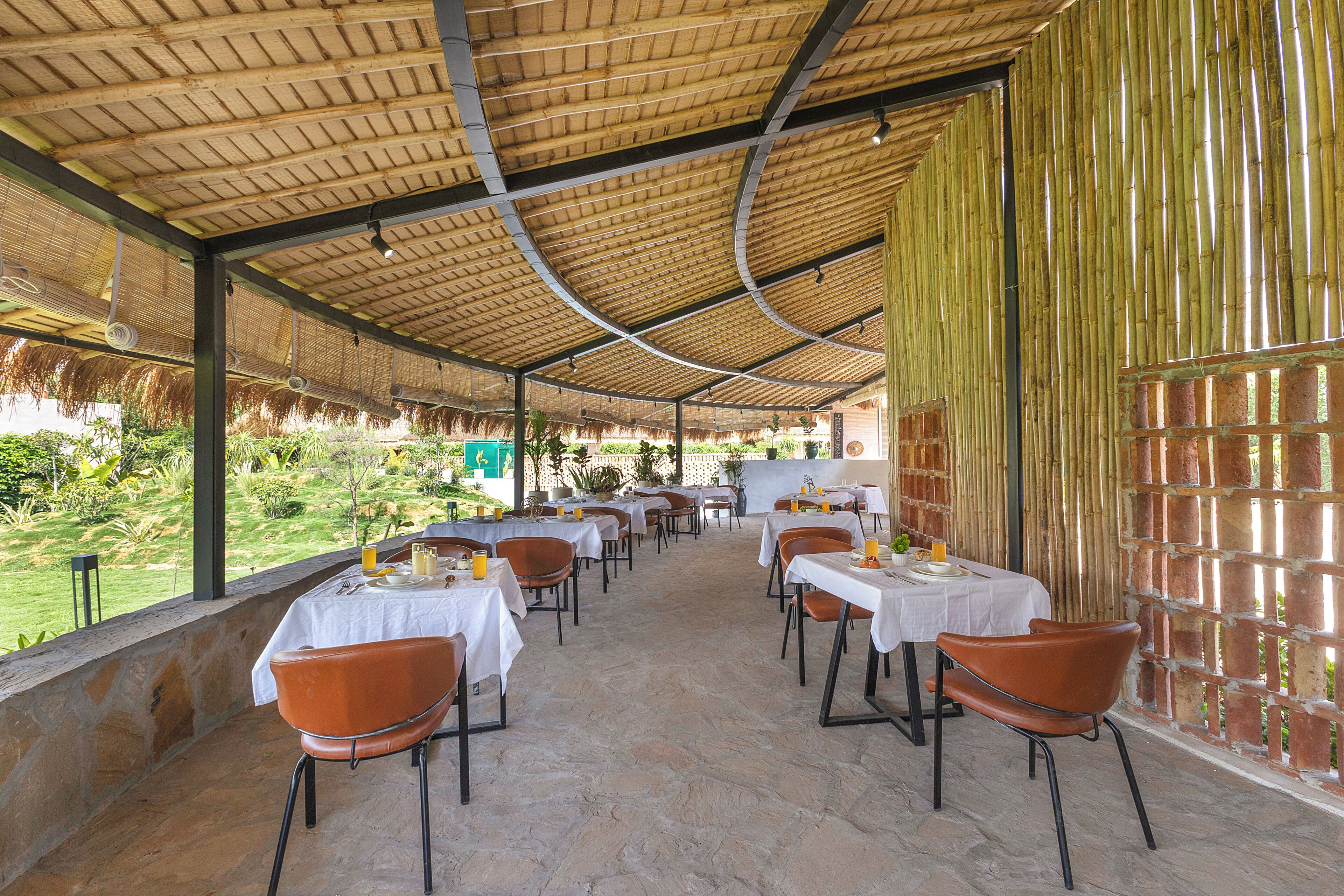 An outdoor dining area with tables and chairs, covered by a bamboo roof, and a stone floor.