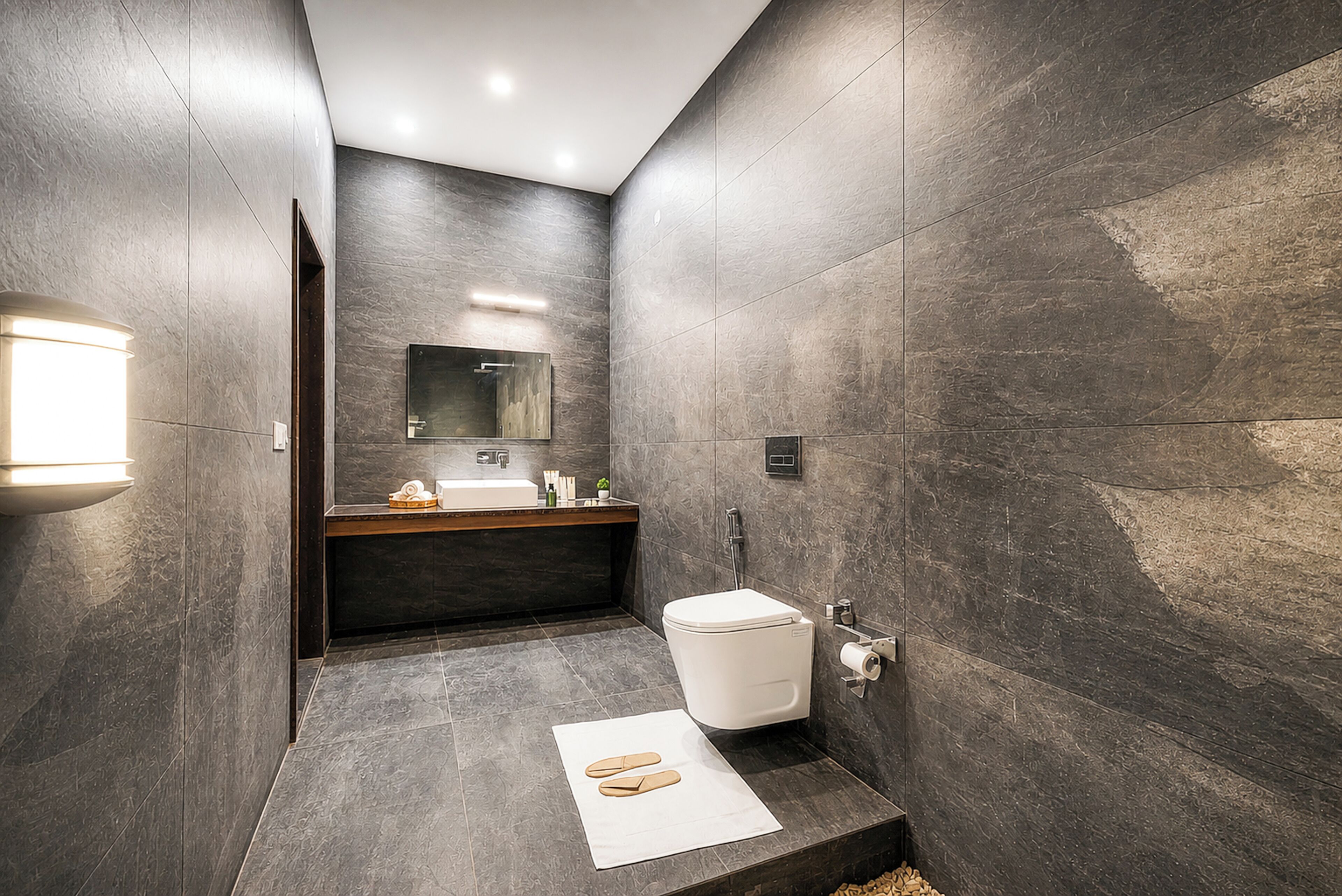 A modern bathroom with dark stone walls, a white toilet, a sink with a mirror above it, and a towel on the floor.