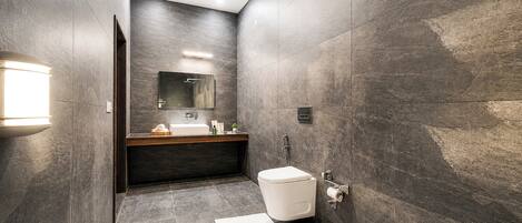 A modern bathroom with dark stone walls, a white toilet, a sink with a mirror above it, and a towel on the floor.