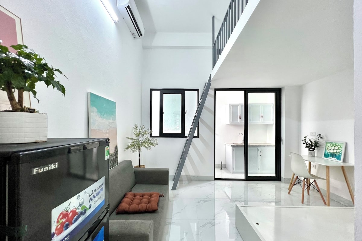 A modern living room with a flat-screen TV, a sofa, a potted plant, a staircase, and a kitchen area.