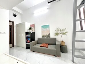 Una sala moderna con un sofá gris, una mesita, una planta en maceta, un refrigerador y un aire acondicionado de pared.