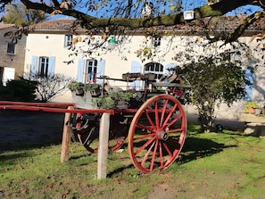 Un bâtiment rustique à l’architecture traditionnelle, un chariot en bois rouge et un petit jardin.