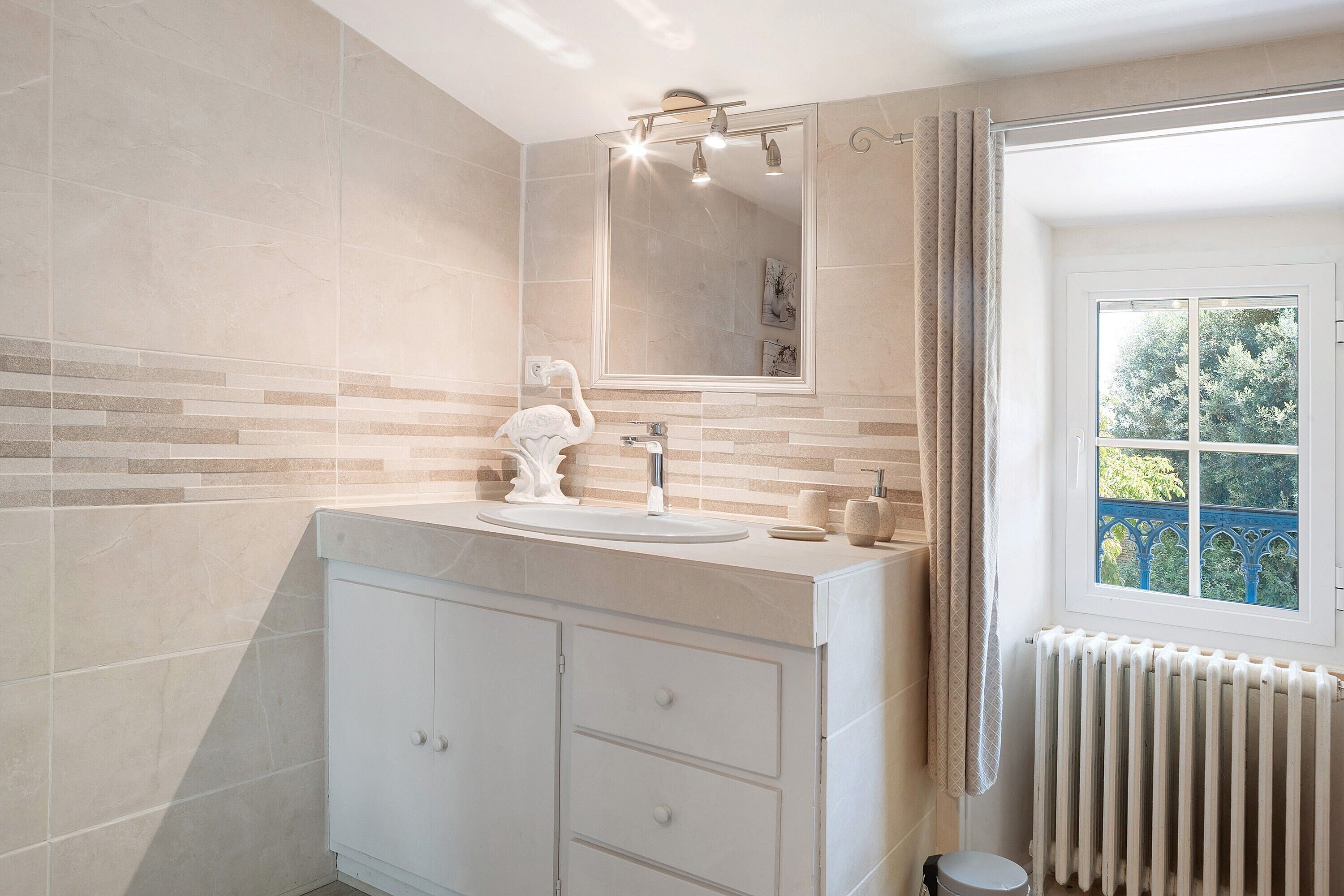 Une salle de bain avec un meuble-lavabo blanc, un grand miroir et une fenêtre donnant sur les arbres.