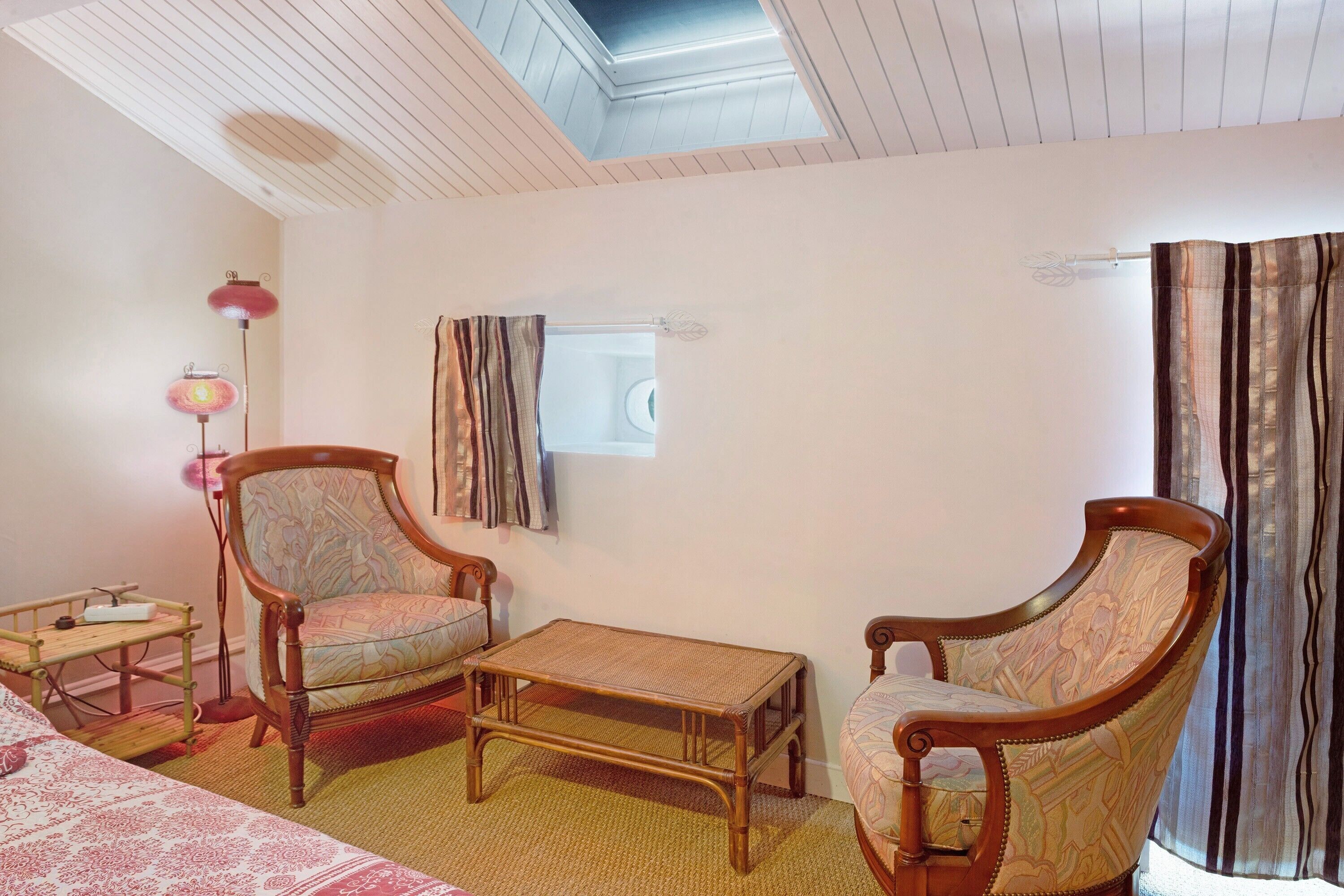 Una habitación con dos sillas de madera, una mesita, una cama, una lámpara y un ventanal con cortinas.