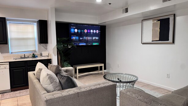 Una sala de estar moderna con un televisor de pantalla plana, sofás grises, una mesa de centro de vidrio y una zona de cocina con un lavabo y armarios.