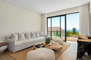 Ein modernes Wohnzimmer mit Sofa, Couchtisch und Hocker, ein Essbereich mit Tisch und eine große Schiebeglasdoor, die auf einen Balkon mit Blick führt.