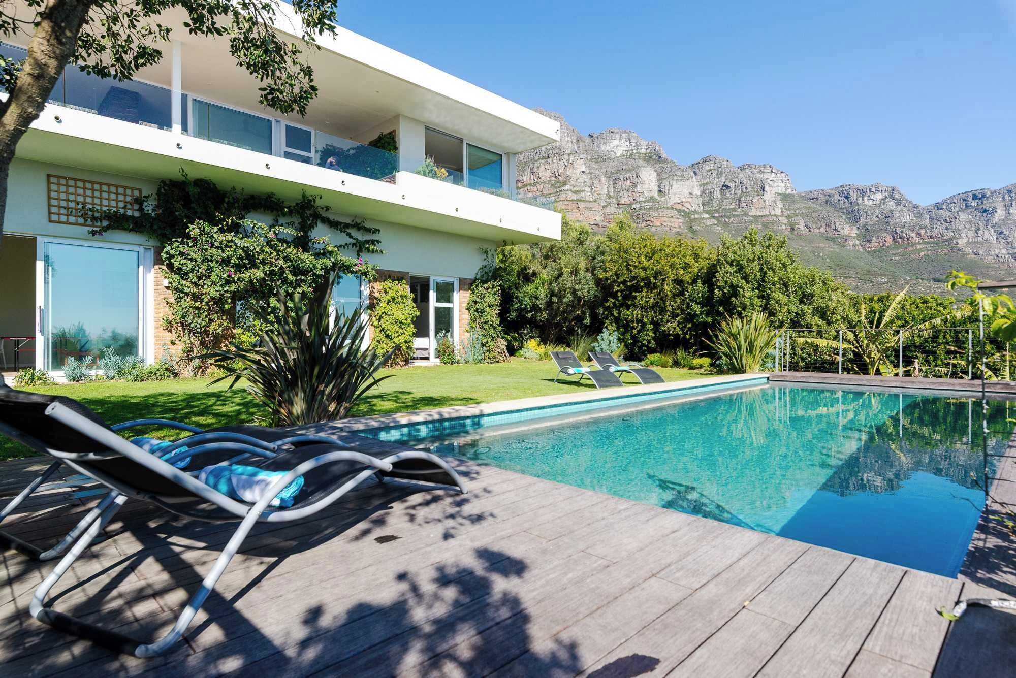 A modern two-story house with a swimming pool, lounge chairs, and a mountain backdrop.