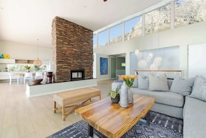 A modern living room with a stone fireplace, a sofa, a coffee table, and a dining area with a bench. - Golden View Villa | The Ultimate Sunset Escape (Cape Town)