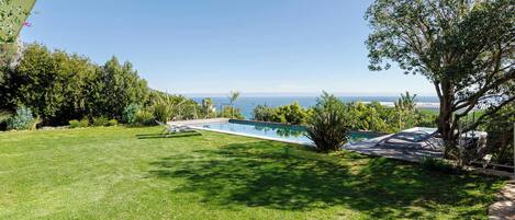 Una piscina con vista al mar, rodeada por un césped y árboles.