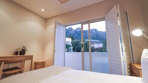 A bedroom with a bed, a desk, a chair, a lamp, and a view of mountains through a sliding door. - Skyloop Villa | Luxury Living for the Whole Family (Cape Town)