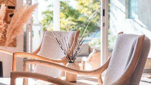 A modern interior with a seating area featuring a wooden chair and a cushioned chair, a vase with dried plants, and a view of greenery outside. - Skyloop Villa | Luxury Living for the Whole Family (Cape Town)