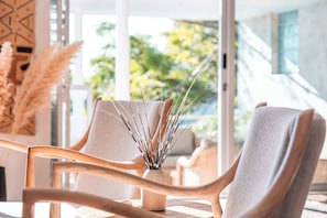 Ein modernes Interieur mit einer Sitzecke, bestehend aus einem Holzstuhl und einem gepolsterten Sessel, einer Vase mit Trockenpflanzen und Blick ins Grüne.