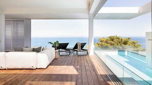 Ein moderner Balkon mit Holzboden, einer weißen Couch, zwei Loungestühlen und einem Glasgeländer mit Blick auf einen Pool und das Meer.