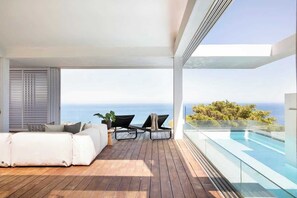Ein moderner Balkon mit Holzboden, einer weißen Couch, zwei Loungestühlen und einem Glasgeländer mit Blick auf einen Pool und das Meer.