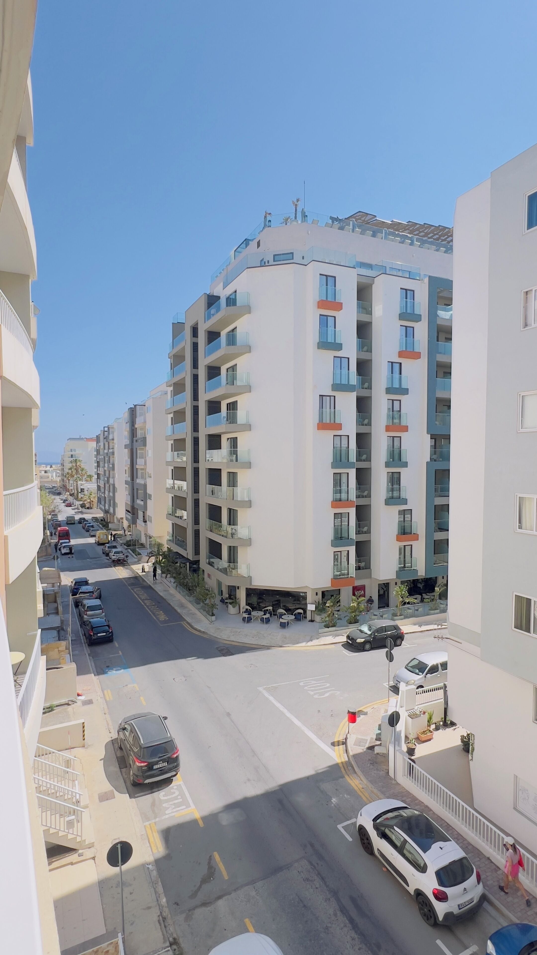 Um edifício residencial com várias andares, varandas, uma rua com carros estacionados e céu claro.