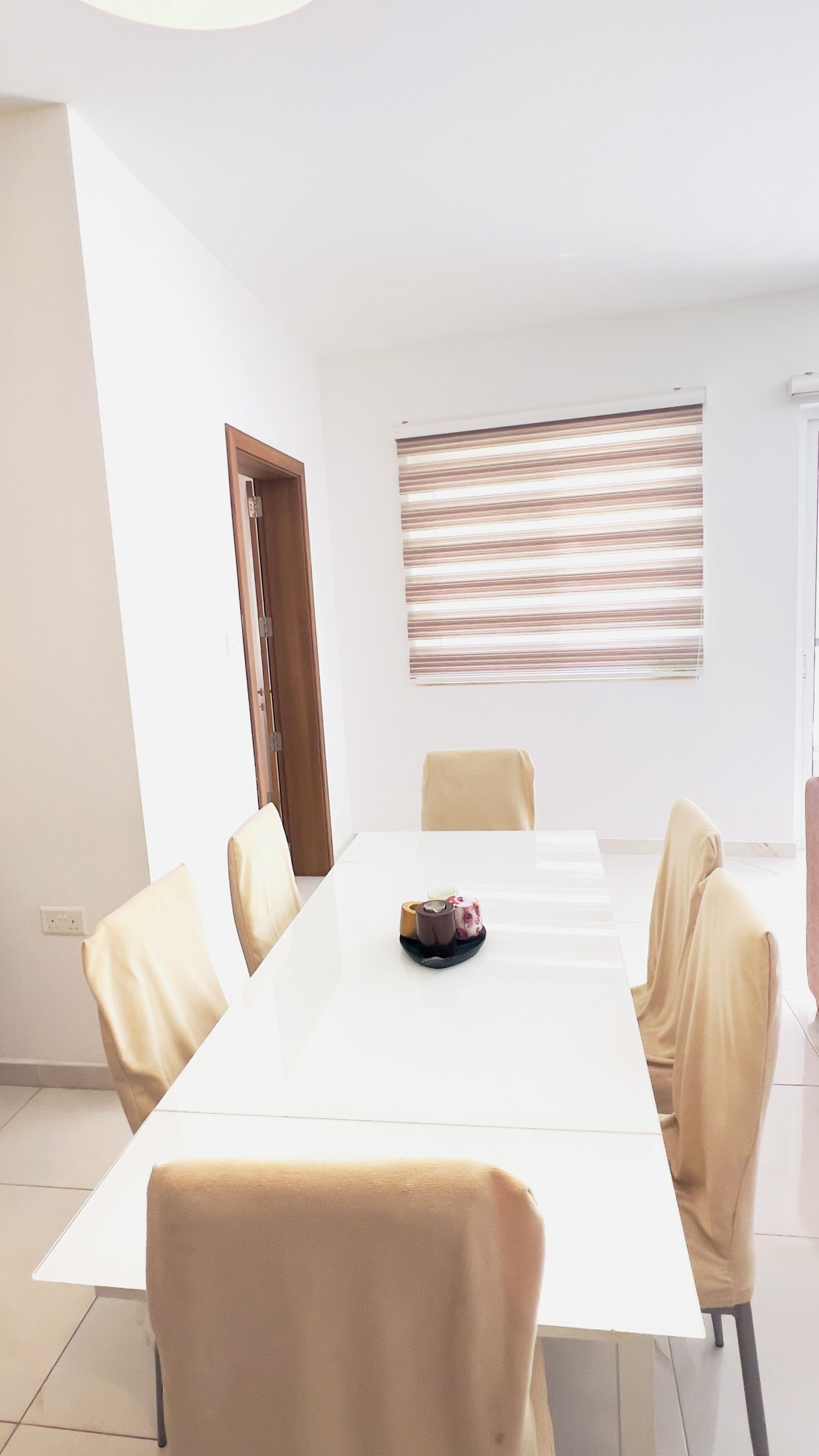 Ett konferensrum med ett långt vitt bord, beige stolar, ett litet bord med en tårta, ett fönster med persienner och en dörr.