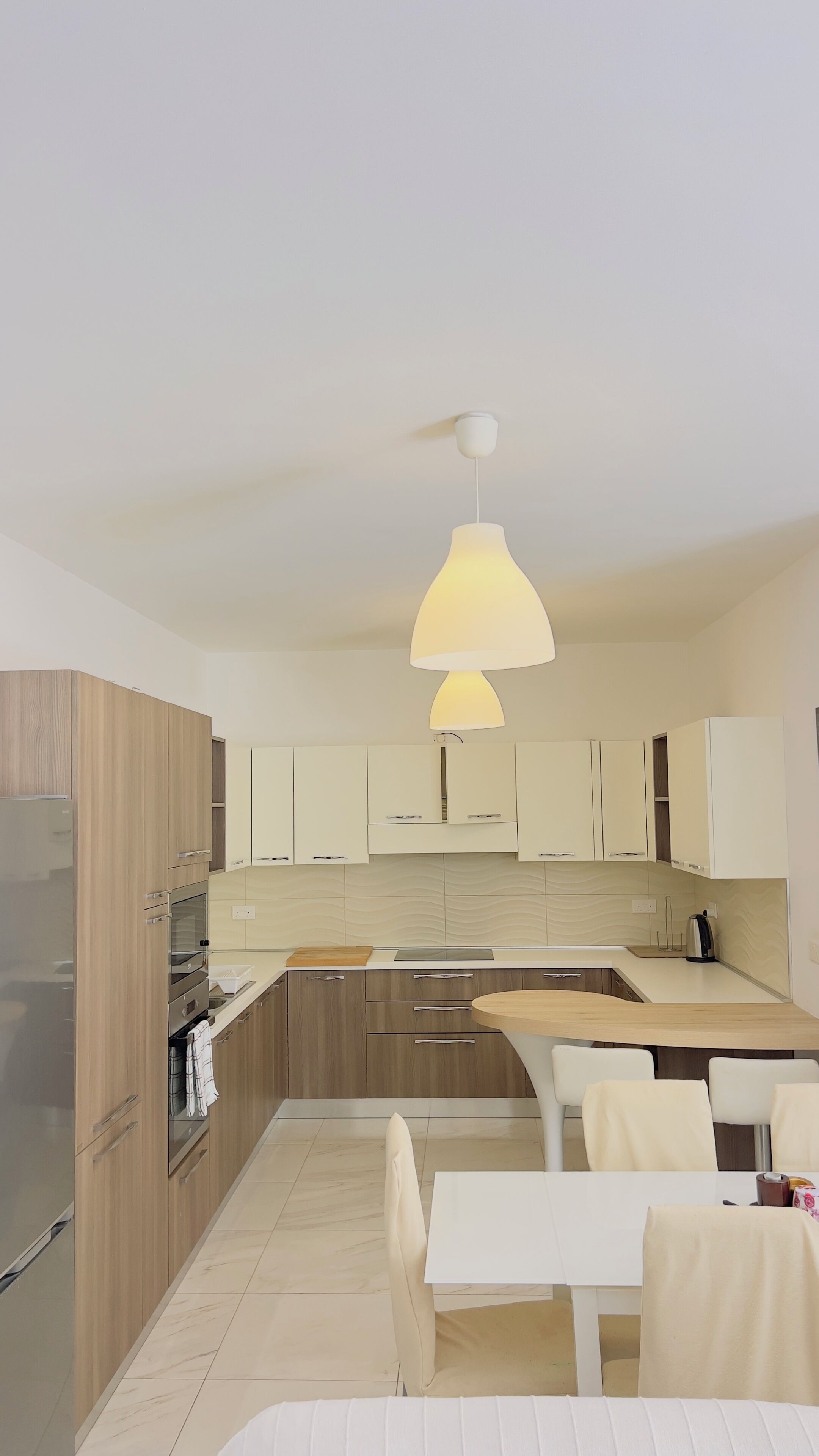 A modern kitchen with wooden cabinets, a white countertop, a built-in oven, and a dining area with a white table and chairs.