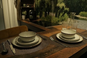 Una mesa de comedor con platos, tazones, cubiertos y vasos, con vista a árboles y una fuente a través de un ventanal.