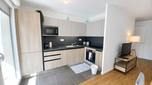 Una cocina moderna con microondas, horno y lavamanos, una pequeña mesa de madera con televisor, y un ventanal con persianas.
