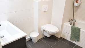 Un baño con lavamanos, inodoro y bañera blancos. Hay un perchero con una toalla, un cesto de basura blanco y una zona de ducha.