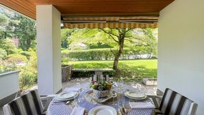 Een overdekt terras met eettafel voor vier personen, omringd door stoelen, en uitzicht op een tuin met een boom en hagen.