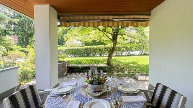 A covered outdoor dining area with a table set for four, surrounded by chairs, and a view of a garden with a tree and hedges. - Villa with a garden, 15 minutes from Como; exclusive residence with a pool and tennis court (Lombardia)