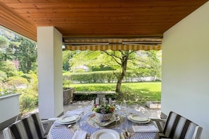 Ein überdachter Essbereich im Freien mit Tisch für vier Personen, umgeben von Stühlen, und Blick auf einen Garten mit Baum und Hecken.
