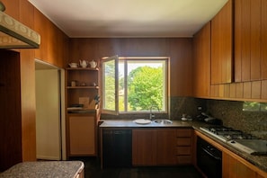 A compact kitchen with wooden cabinets, a built-in dishwasher, a window with a view of greenery, and a countertop with a sink and stove.