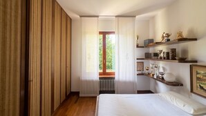 A bedroom with a bed, a window with curtains, a radiator, and shelves with decorative items.