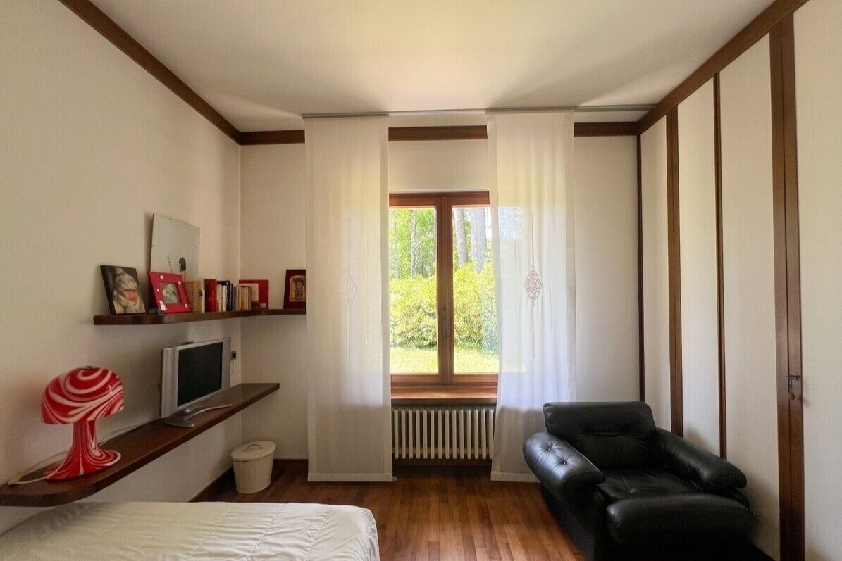 Ein Zimmer mit einem Bett, einem schwarzen Ledersofa, einem Fernseher auf einem Regal, einem Fenster mit Vorhängen und einem Heizkörper.