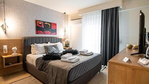 A modern hotel room with a large bed, a desk, a nightstand, a painting on the wall, and a window with curtains. - Alexandra Residence Seaside (ALEXANDROUPOLIS)