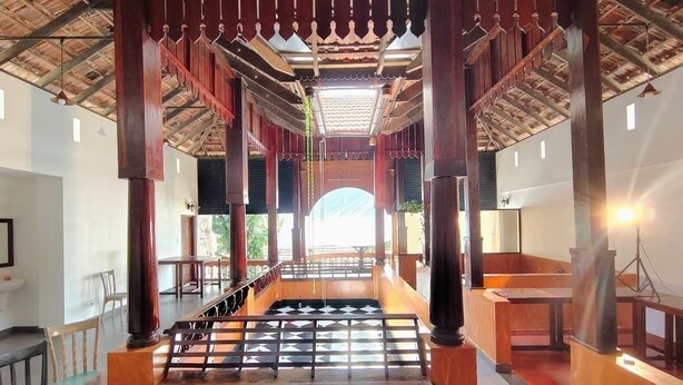 A spacious indoor area with wooden pillars, a central seating area, and a view through a large archway. - Samudra Heritage (Kochi)