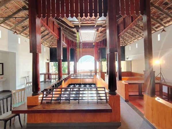 A spacious indoor area with wooden pillars, a central seating area, and a view through a large archway.