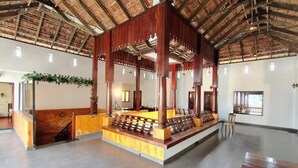 A spacious hall with a high ceiling and wooden beams, a central wooden structure with a bench, and a staircase leading to an upper level. - Samudra Heritage (Kochi)