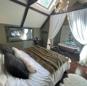 Un dormitorio con cama, espejo, candelabro, ventana con cortinas y una bañera.