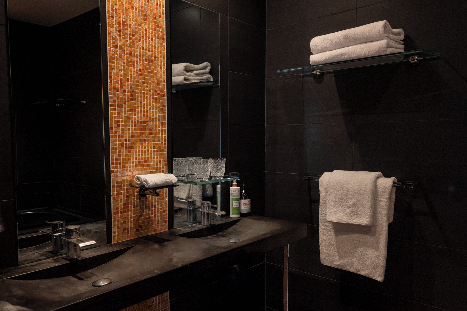 A modern bathroom with a dark tile wall, a glass shelf with a folded towel, a glass sink, and a mirror with a mosaic tile backsplash.