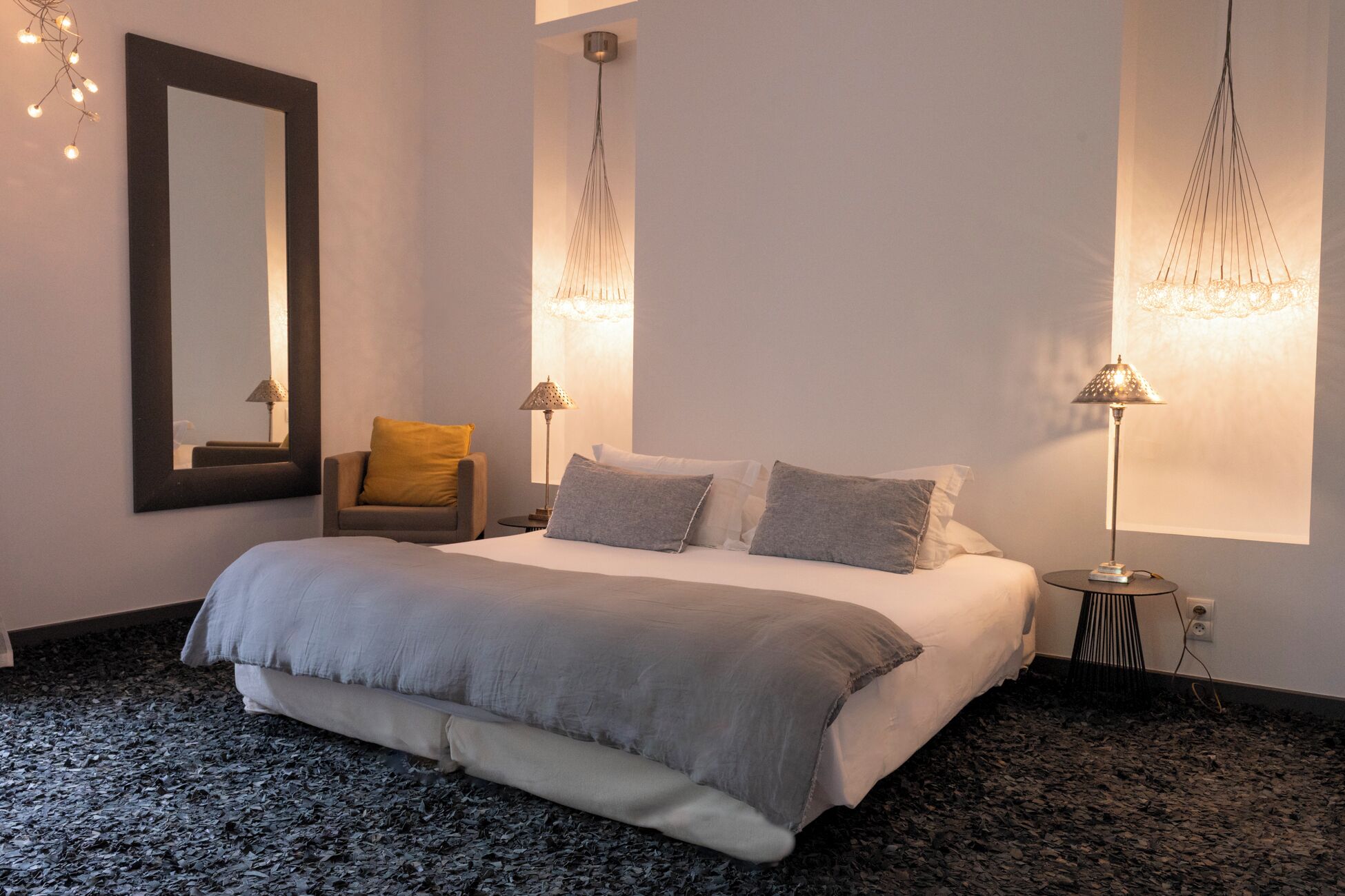 Un dormitorio con una cama, una silla, dos lámparas de noche y un espejo en la pared.