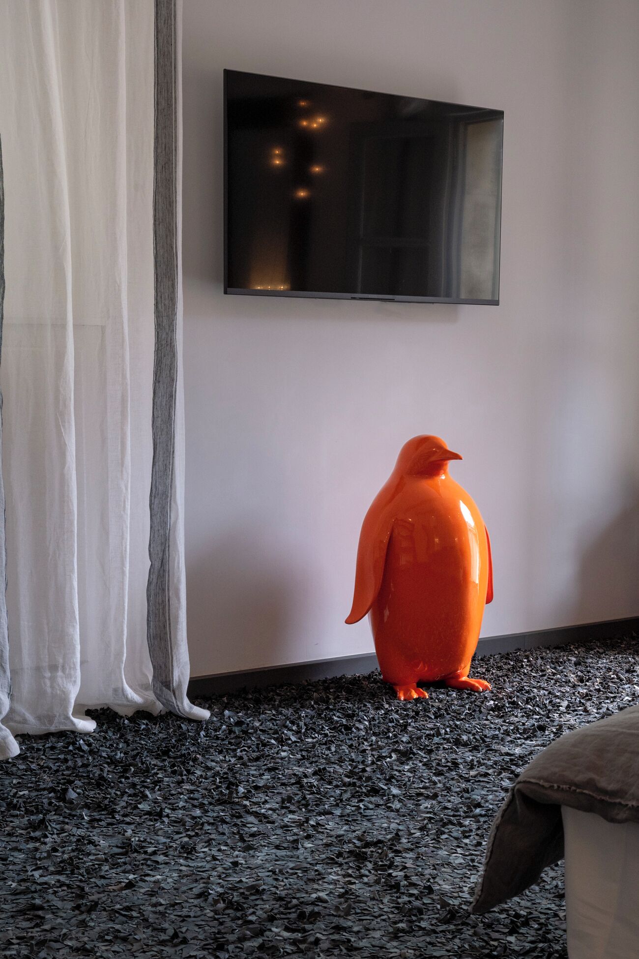A room with a large flat-screen TV mounted on the wall, a white curtain, and an orange sculpture on a dark floor.