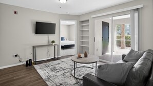 Een moderne woonkamer met een flatscreen-tv, een ronde salontafel, een bank, een grote schuifpui naar een balkon en een zichtbare slaapkamer met een bed.