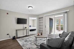 Ett modernt vardagsrum med en platt-TV, ett runt soffbord, en soffa, en stor skjutdörr i glas som leder ut till en balkong och ett synligt sovrum med en säng.