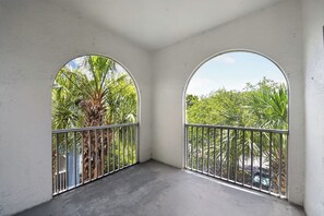 Un balcon doté de deux fenêtres en arc de cercle, d’une balustrade en métal et offrant une vue sur des palmiers et une végétation luxuriante.