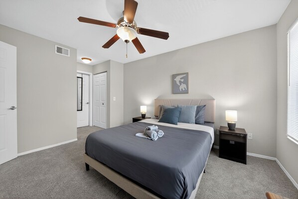 Ein Schlafzimmer mit einem Bett, einem Deckenventilator, einem Nachttisch mit Lampe, einer Tür und einem gerahmten Bild an der Wand.