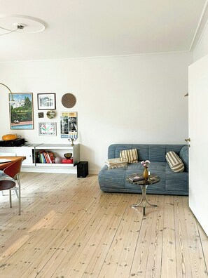 Un salón con un sofá azul, una mesa de centro, suelo de madera y una pared blanca con marcos de cuadros.