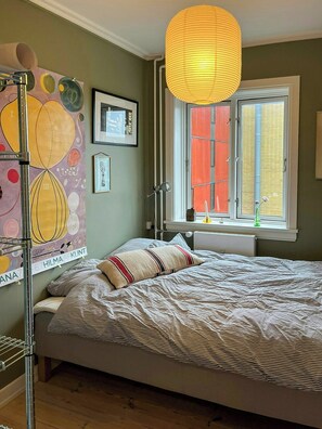 Un dormitorio con cama, ventana con persianas, lámpara colgante y cuadros en la pared.