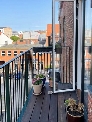 Un balcón con plantas en macetas, baranda de metal y vista de edificios al fondo.