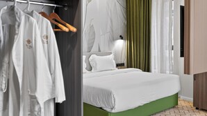 Un dormitorio con estructura de cama verde, cubrecama blanco, mesita de noche con lámpara y armario con ropa colgada.