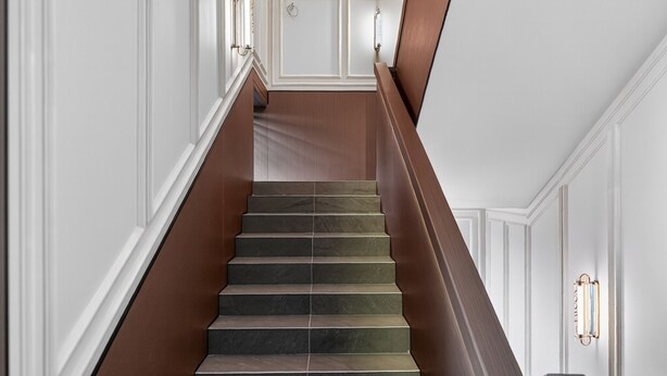 Una escalera con barandilla de madera oscura y peldaños, paredes blancas con paneles y luminarias fijadas a la pared.