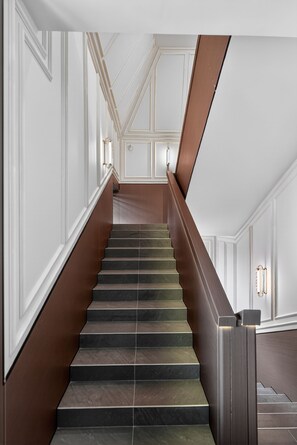 Una escalera con barandilla de madera oscura y peldaños, paredes blancas con paneles y luminarias fijadas a la pared.