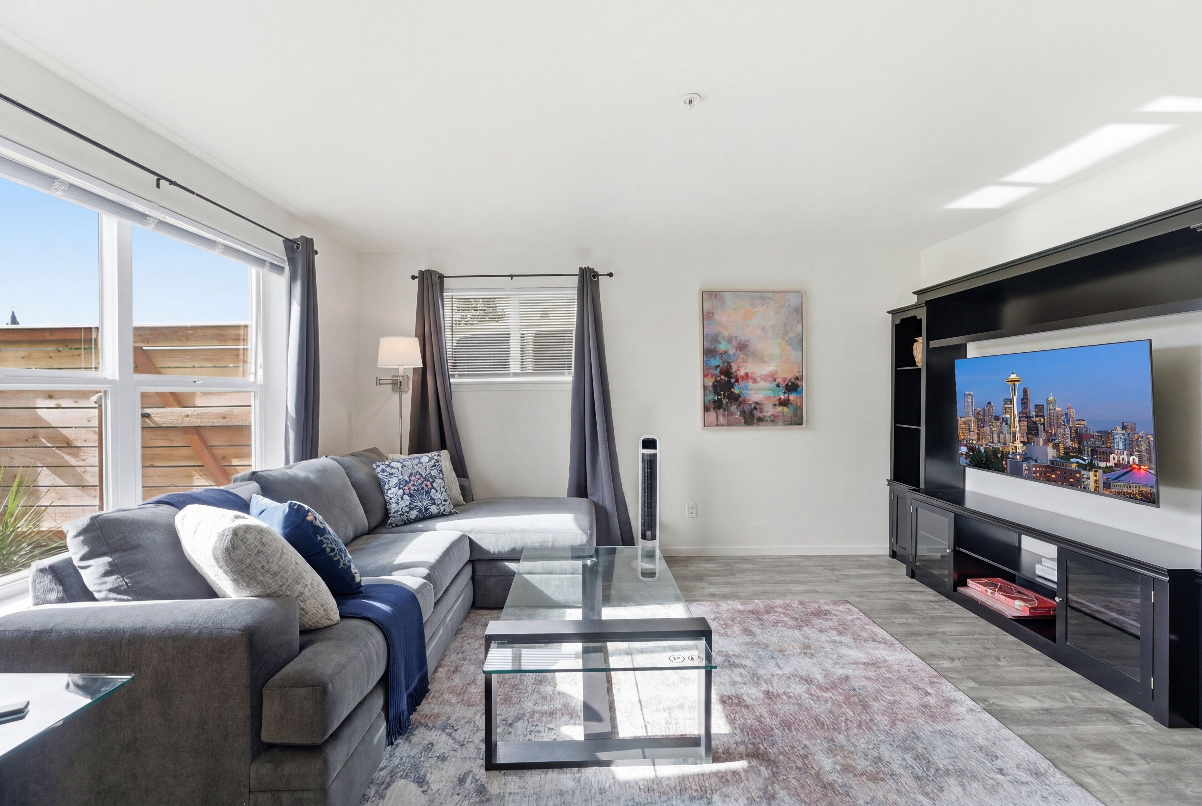 A modern living room with a sectional sofa, a glass coffee table, a flat-screen TV, and a cityscape painting.