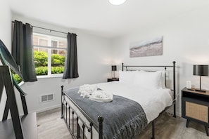 Ein Schlafzimmer mit Bett, einem Fenster mit Vorhängen, einem Nachttisch mit Lampe und einem Bild an der Wand.